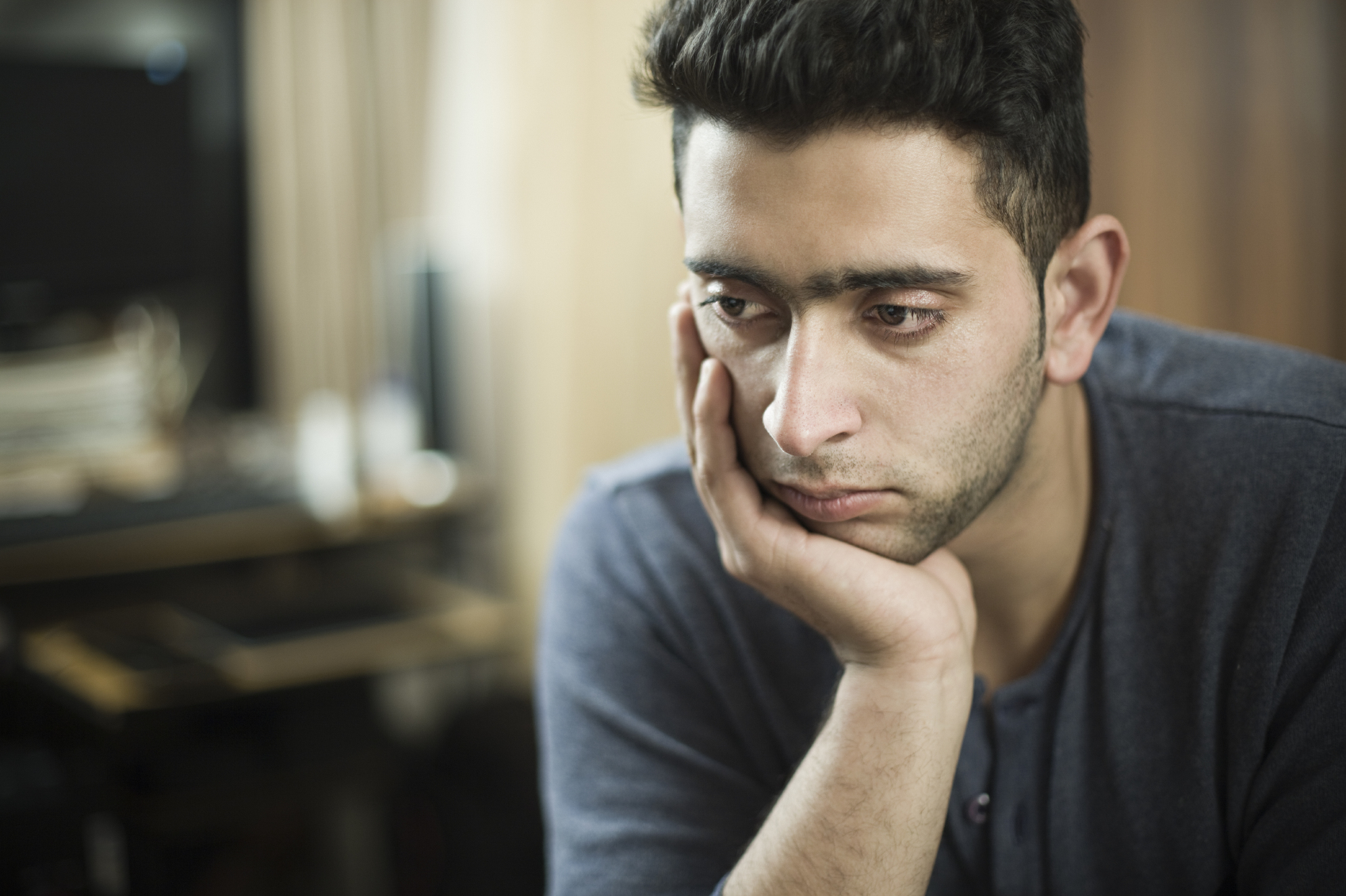 Indoor low key image of a serene young man thinking by resting his head on hand and looking away with and blank expression. He is wearing a t-shirt. One person, waist up, horizontal composition with selective focus and copy space.