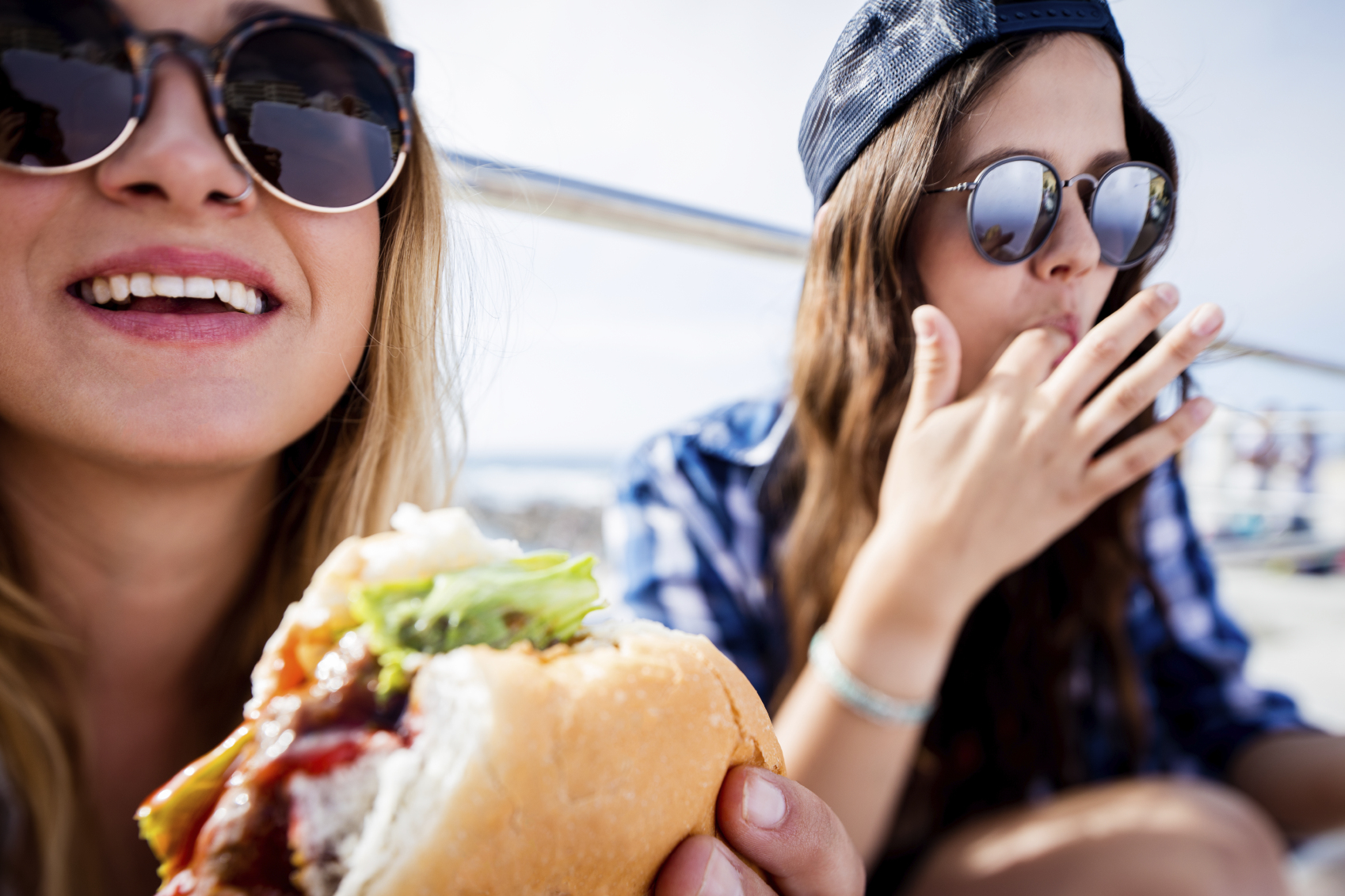 Teenage girl friends wearing hipster sunglasses sitting outdoors on a summer day eating large delicious hamburgers together