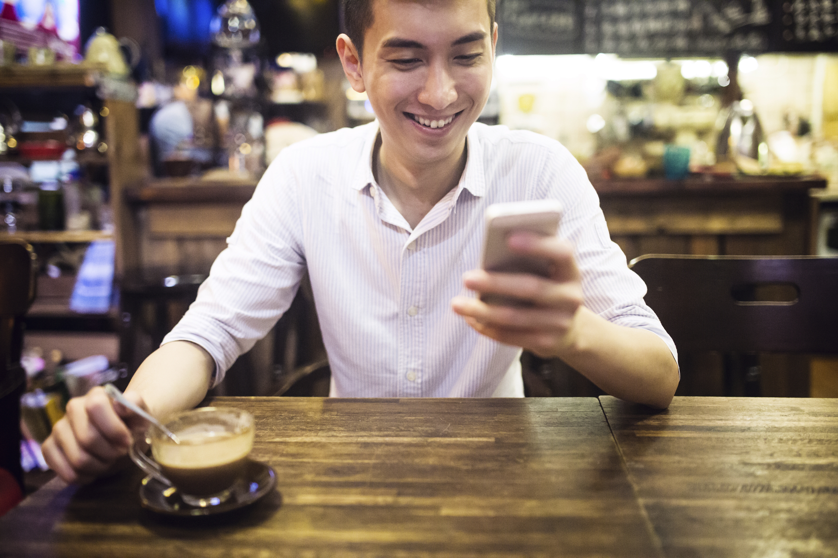A young Chinese man enjoys a cappuccino in a coffee and tea shop in Hong Kong.  He smiles while sending a text on his cell phone.  Horizontal image.