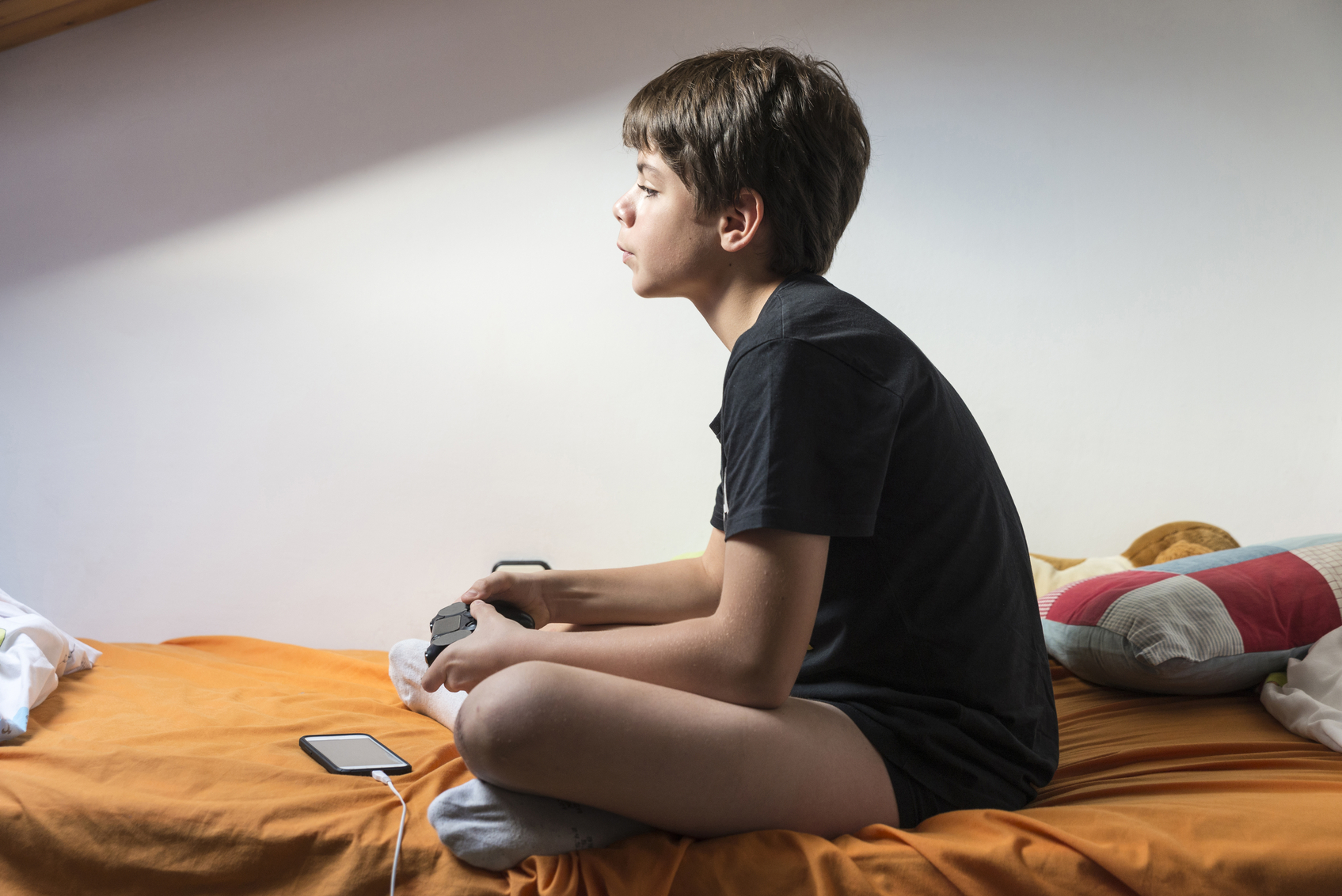 Young boy playing video games sitting on the bed in her room while charging the phone