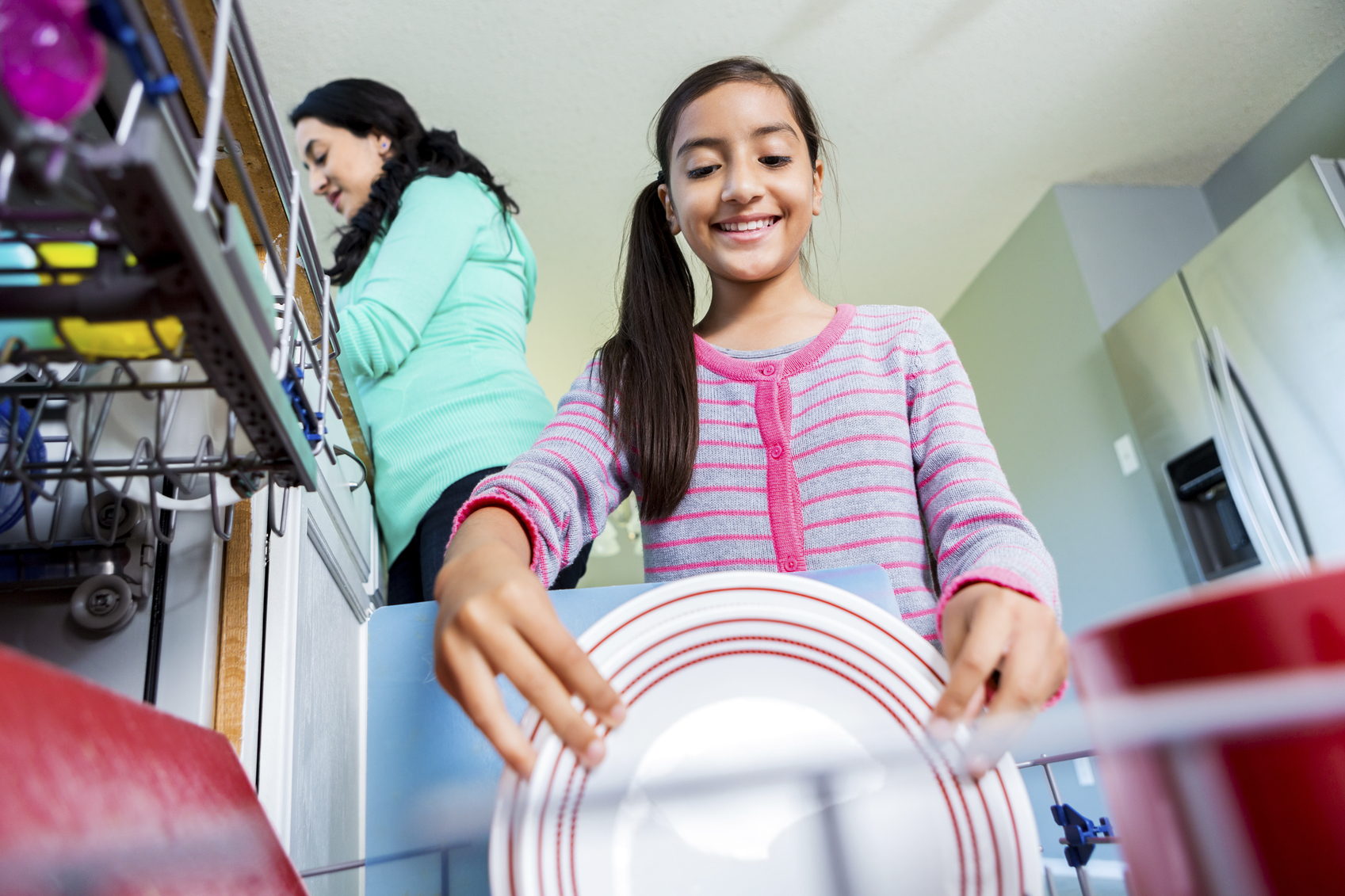 Cute pre-teen Hispanic girl smiles as she loads a plate into the dishwasher. Her mom is rinsing off the dishes in the background. Photo shot from low angle view point.